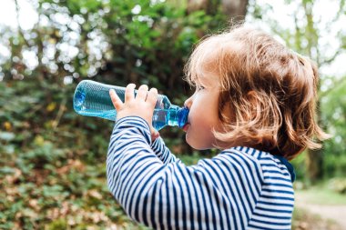 Küçük sarı saçlı kız doğadaki bir gezi sırasında plastik şişeden su içiyor.