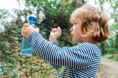Sarışın, küçük bir kız doğada yürürken su dolu plastik bir şişeye bakıyor.