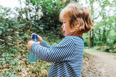 Küçük sarı saçlı kız doğadaki bir gezi sırasında plastik su şişesini kapatıyor.
