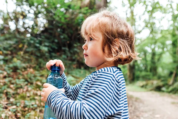 Küçük sarı saçlı kız doğadaki bir gezi sırasında plastik bir su şişesini açıyor.