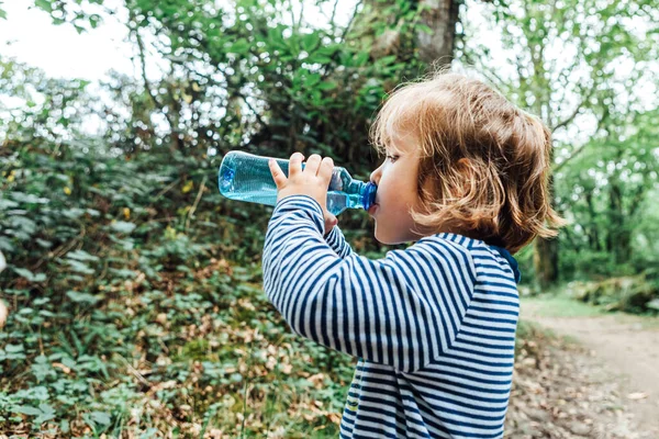 Küçük sarı saçlı kız doğadaki bir gezi sırasında plastik şişeden su içiyor.