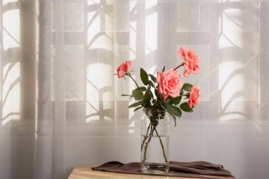 Beyaz perdeli ahşap bir masanın üzerine yerleştirilmiş pembe güller, odanın içinde hüzünlü bir hava ve huzurlu bir his yaratır..