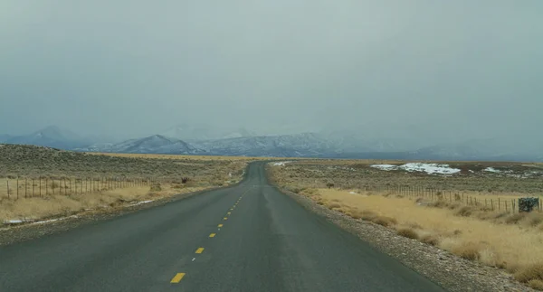 Önünde karlı dağ sıraları ve arkaplanda bulutlu gökyüzü olan düz bir yol.