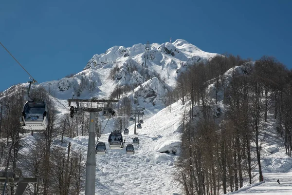 Montañas nevadas de la estación de esquí Krasnaya Polyana en Sochi en el que el teleférico con los turistas. Cielo azul claro en el bosque montañoso. Temporada turística de esquí y snowboard — Foto de Stock