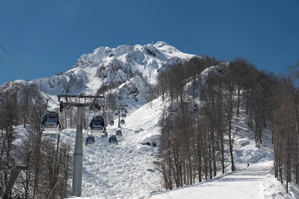 Montañas nevadas de la estación de esquí Krasnaya Polyana en Sochi en el que el teleférico con los turistas. Cielo azul claro en el bosque montañoso. Temporada turística de esquí y snowboard — Foto de Stock