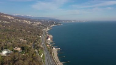Havadan video çekimi. Soçi'nin Karadeniz kıyıları bir yükseklikten. Kamera kıyıdan hareket ediyor. Kıyı şeridi. Sahil boyunca uzanan yol. Krasnaya Polyana Dağları.
