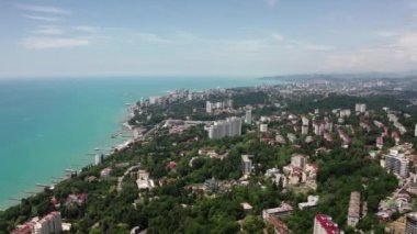 Havadan video çekimi. Soçi şehrinin zirvesinden panoramik manzara. Yeşil ağaçlar ve deniz deki dağlar. Yerleşim alanı. Sahilde ki yüksek evler. Mavi ve berrak gökyüzü.
