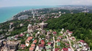 Havadan video çekimi. Soçi şehrinin zirvesinden panoramik manzara. Yeşil ağaçlar ve deniz deki dağlar. Yerleşim alanı. Sahilde ki yüksek evler. Mavi ve berrak gökyüzü.