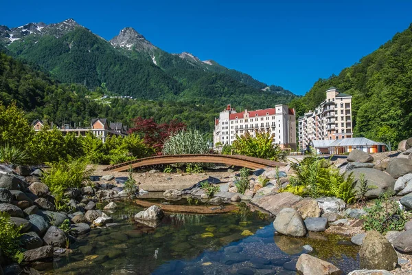 Rosa Khutor. Krasnaya Polyana. Soçi. Yüksek dağlar. Yeşil ağaçlar. Mavi gökyüzü. Burada kal. Doğa. Panoramik manzara. Göl. Nehrin üzerinde tahta bir köprü. - Evet. Yüksek kalite fotoğraf
