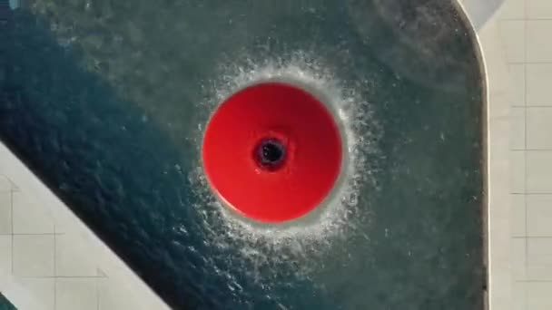 ドローンは水を噴き出す赤い噴水の上でゆっくりと上昇します 噴水の周りの半円にテーブルが立っている — ストック動画