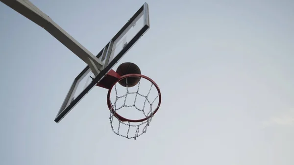 Bola de basquete marrom cair através da cesta com rede de corrente de metal no dia ensolarado de verão no fundo do céu azul claro. Conceito de campo de jogo urbano moderno . — Fotografia de Stock