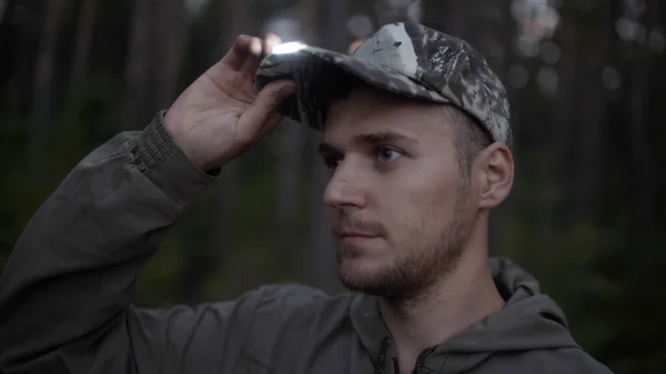 Portrait of handsome young man hunter or tourist. Man puts on his head cap with diode flashlight on visor in evening.