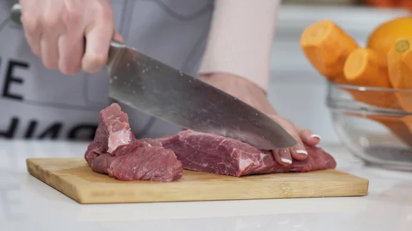 Woman cutting raw meat on small pieces with kitchen knife cutting board. Female cutting red beef filet on wooden board, holding knife in hand. Housewife is cooking at home interior. Concept: food