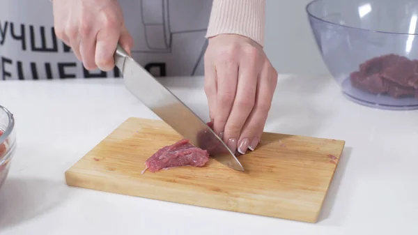 Woman cutting raw meat on small pieces with kitchen knife cutting board. Female cutting red beef filet on wooden board, holding knife in hand. Housewife is cooking at home interior. Concept: food