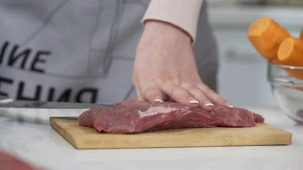 Woman cutting raw meat on small pieces with kitchen knife cutting board. Female cutting red beef filet on wooden board, holding knife in hand. Housewife is cooking at home interior. Concept: food