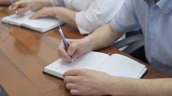 Male hand holding pen and writing to do list or business data with pen at notebook dairy. Hand writing business report, make note on paperwork document on office desk in corporate meeting room.