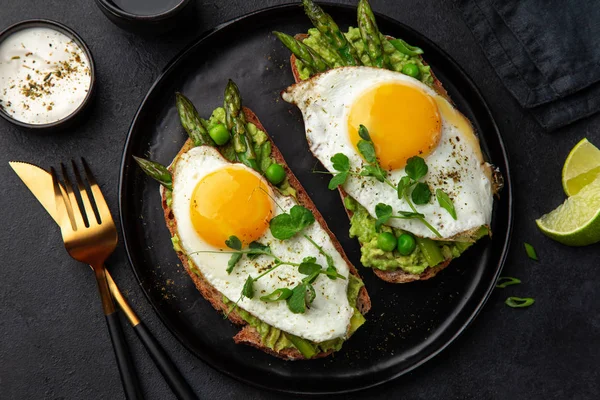 Tostadas con aguacate, espárragos y huevo frito en plato negro —  Fotos de Stock