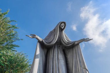 Meryem Ana heykeli bronz, mavi gökyüzü arka plan beyaz bulutlar ve ağaç, uzanmış kollarıyla.