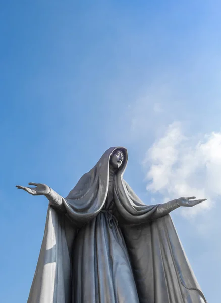 Statue of Virgin Mary in bronze, in a blue sky background with white clouds, with outstretched arms.