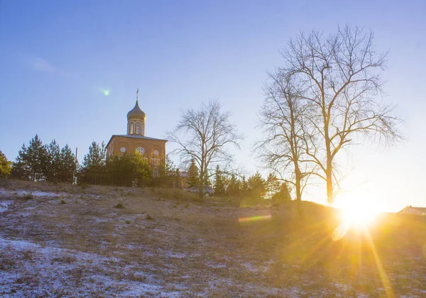 Iglesia ortodoxa en una colina cerca del bosque. Paisaje de atardecer de invierno con cielo azul claro y rayos de sol — Foto de Stock