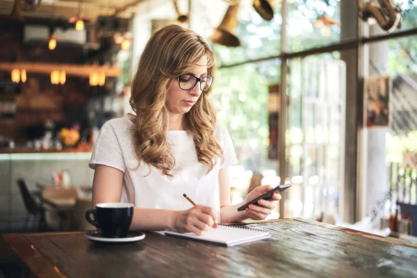 年轻金发女郎使用智能手机和在咖啡馆做笔记 — 图库照片