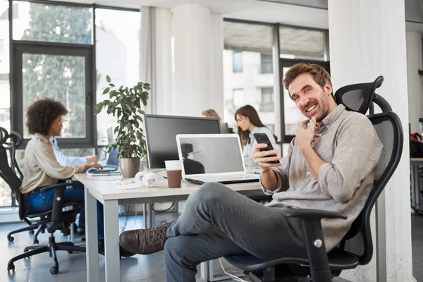 CEO with smile sitting in his chair and using smart phone. Office interior, in background employees working.