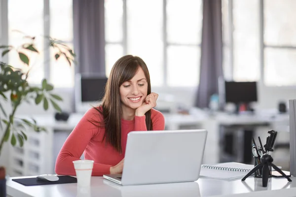 Красивая улыбающаяся кавказская деловая женщина сидит в современном офисе и пользуется ноутбуком . — стоковое фото