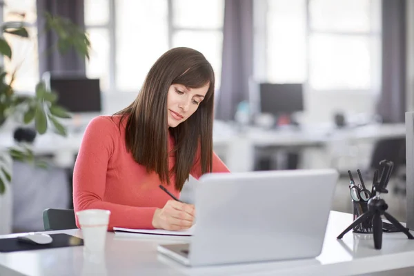 Encantadora mujer de negocios caucásica sentada en la oficina moderna y tomando notas en un cuaderno. Delante de su portátil . — Foto de Stock