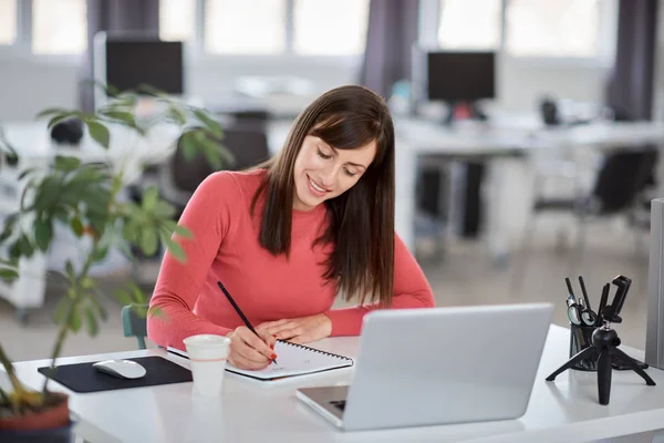 Charming Caucasian businesswoman sitting in moderna office and taking notes in notebook. In front of her laptop. — Stock Photo, Image