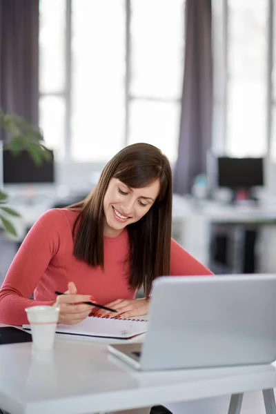 Encantadora mujer de negocios caucásica sentada en una oficina moderna y tomando notas en un cuaderno. Delante de su portátil . — Foto de Stock