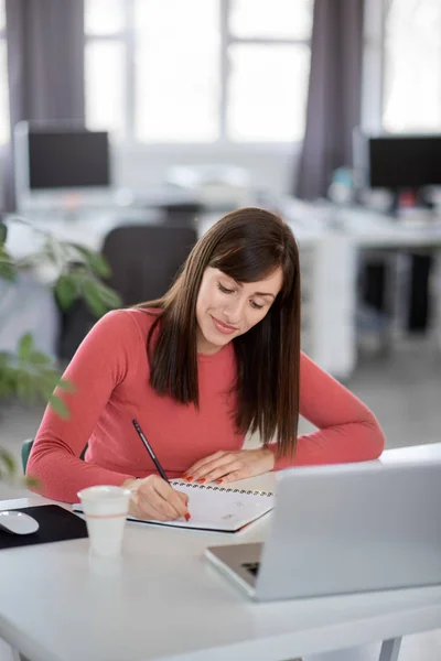 Die charmante kaukasische Geschäftsfrau sitzt in einem modernen Büro und macht sich Notizen in einem Notizbuch. vor ihrem Laptop. — Stockfoto