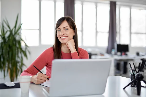 Красивая улыбающаяся кавказская деловая женщина сидит в современном офисе и пользуется ноутбуком . — стоковое фото
