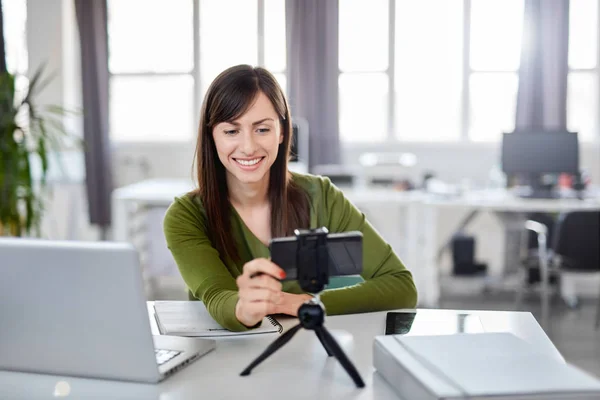 Charming Caucasian brunette in green outfit adjusting smart phone for video call. In front of her laptop, office interior. — Stock Photo, Image