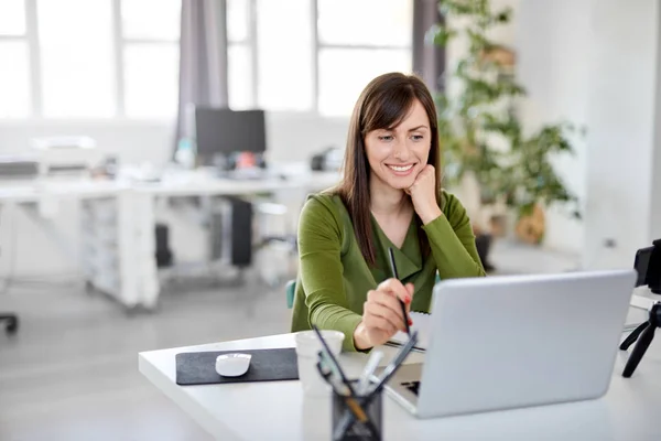 Giovane caucasico occupato bruna sorridente e in possesso di matita mentre seduto in ufficio e guardando il computer portatile . — Foto Stock