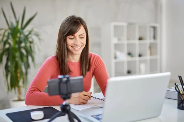 Charming Caucasian businesswoman sitting in modern office and taking notes in notebook. In front of her laptop. — Stock Photo, Image