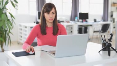 Young beautiful Caucasian businesswoman with brown hair sitting in office and using laptop.