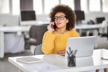 Sevimli karışık ırk iş kadını modern ofiste oturan ve akıllı telefon kullanarak rahat giyinmiş.