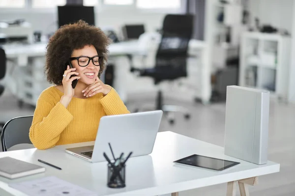 Encantadora mujer de negocios de raza mixta vestida casual usando un teléfono inteligente mientras está sentada en una oficina moderna . — Foto de Stock