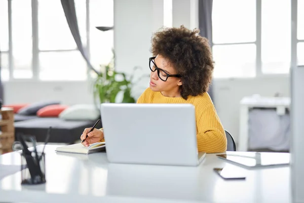 Attraktiv blandad ras affärskvinna klädd casual anteckningar i anteckningsboken när du sitter i moderna kontor. På skrivbord laptop, surfplatta och Smart Phone. — Stockfoto