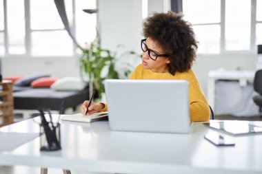 Modern ofiste otururken dizüstü notlar alarak rahat giyinmiş çekici karışık ırk iş kadını. Masa başında dizüstü bilgisayar, tablet ve akıllı telefon.