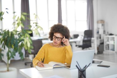 Modern ofiste otururken dizüstü notlar alarak rahat giyinmiş çekici karışık ırk iş kadını. Masa başında dizüstü bilgisayar, tablet ve akıllı telefon.