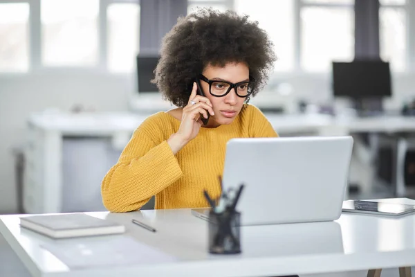 Schöne gemischte Geschäftsfrau, die lässig gekleidet ist und Laptop und Smartphone gleichzeitig benutzt, während sie im modernen Büro sitzt. — Stockfoto