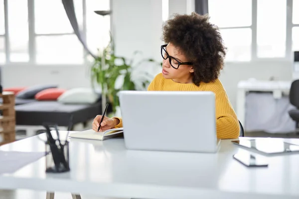 O femeie de afaceri atractivă de rasă mixtă îmbrăcată ocazional luând notițe în caiet în timp ce stătea în biroul modern. Pe laptop, tabletă și telefon inteligent . — Fotografie, imagine de stoc