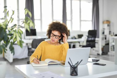 Modern ofiste otururken dizüstü notlar alarak rahat giyinmiş çekici karışık ırk iş kadını. Masa başında dizüstü bilgisayar, tablet ve akıllı telefon.