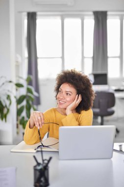 Büyüleyici karışık ırk iş kadını rahat gözlük tutarak giyinmiş ve modern ofiste otururken uzağa bakıyor.