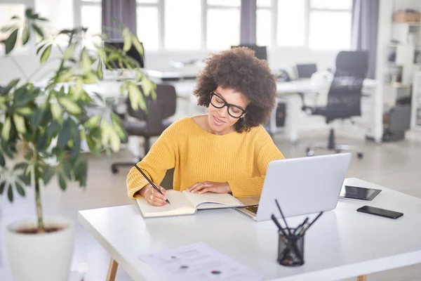 O femeie de afaceri atractivă de rasă mixtă îmbrăcată ocazional luând notițe în caiet în timp ce stătea în biroul modern. Pe laptop, tabletă și telefon inteligent . — Fotografie, imagine de stoc