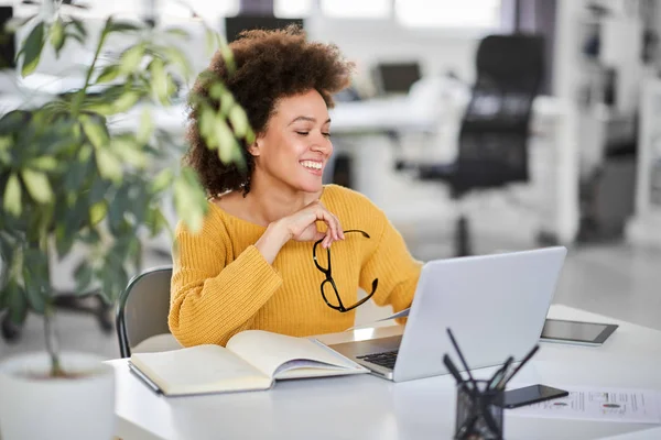 Sorrindo mulher de negócios de raça mista segurando óculos e olhando para o laptop enquanto sentado no escritório moderno . — Fotografia de Stock