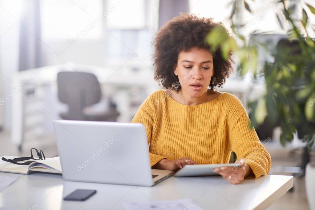 Serious mixed race businesswoman dressed  casual sitting in office and using tablet.
