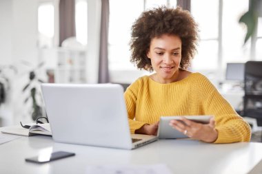 Ciddi karışık ırk iş kadını ofiste rahat oturan ve tablet kullanarak giyinmiş.