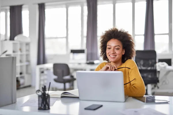 Güzel gülümseyen karışık ırk iş kadını ofiste oturan ve dizüstü bilgisayar kullanarak rahat giyinmiş. — Stok fotoğraf